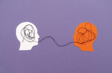 Two Paper Silhouette Heads Connected by Black Thread Symbolizing Mental Connection on Purple Background