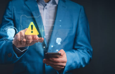 Sinais de alerta na recuperação de crédito empresarial Profissional aponta para um ícone de alerta digital enquanto segura um celular, representando atenção a riscos e falhas na recuperação de crédito empresarial.