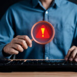 Profissional utiliza lupa com símbolo de alerta diante de um computador, representando a identificação de sinais de fraude à execução.