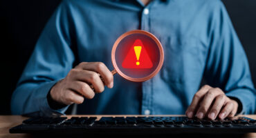 Profissional utiliza lupa com símbolo de alerta diante de um computador, representando a identificação de sinais de fraude à execução.