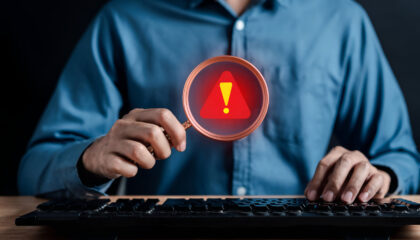 Profissional utiliza lupa com símbolo de alerta diante de um computador, representando a identificação de sinais de fraude à execução.