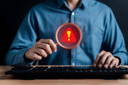 Profissional utiliza lupa com símbolo de alerta diante de um computador, representando a identificação de sinais de fraude à execução.