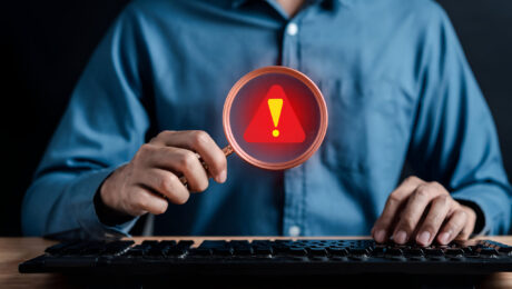 Fraude à execução: identificação de sinais de alerta na recuperação de crédito Profissional utiliza lupa com símbolo de alerta diante de um computador, representando a identificação de sinais de fraude à execução.