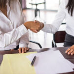 Duas profissionais selando um acordo extrajudicial com aperto de mãos sobre mesa com documentos técnicos e caneta.