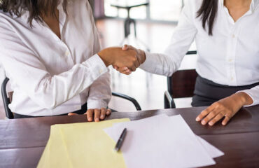 Duas profissionais selando um acordo extrajudicial com aperto de mãos sobre mesa com documentos técnicos e caneta.
