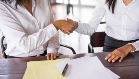 Duas profissionais selando um acordo extrajudicial com aperto de mãos sobre mesa com documentos técnicos e caneta.