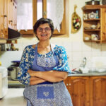 Mulher sorridente de óculos e avental azul com detalhes em patchwork, em pé com os braços cruzados na bancada de uma cozinha rústica com armários de madeira, representando a dona de casa que se dedica ao lar.