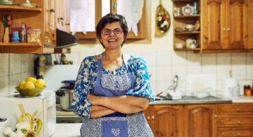 Mulher sorridente de óculos e avental azul com detalhes em patchwork, em pé com os braços cruzados na bancada de uma cozinha rústica com armários de madeira, representando a dona de casa que se dedica ao lar.