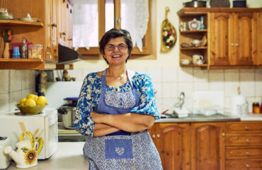 Aposentadoria de dona de casa Mulher sorridente de óculos e avental azul com detalhes em patchwork, em pé com os braços cruzados na bancada de uma cozinha rústica com armários de madeira, representando a dona de casa que se dedica ao lar.