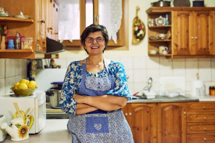 Mulher sorridente de óculos e avental azul com detalhes em patchwork, em pé com os braços cruzados na bancada de uma cozinha rústica com armários de madeira, representando a dona de casa que se dedica ao lar.