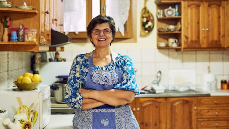 Mulher sorridente de óculos e avental azul com detalhes em patchwork, em pé com os braços cruzados na bancada de uma cozinha rústica com armários de madeira, representando a dona de casa que se dedica ao lar.