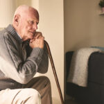Homem idoso de perfil, sentado em uma poltrona, segurando uma bengala de madeira e com expressão pensativa. Ele veste um cardigã cinza e está em uma sala de estar iluminada pela luz do sol.