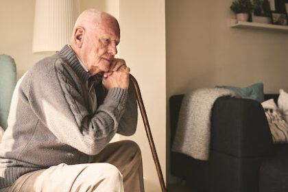 Homem idoso de perfil, sentado em uma poltrona, segurando uma bengala de madeira e com expressão pensativa. Ele veste um cardigã cinza e está em uma sala de estar iluminada pela luz do sol.