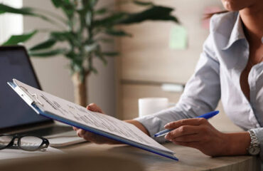 Mulher sentada à mesa de escritório segurando uma prancheta com documentos e uma caneta azul, representando a análise jurídica de dívidas empresariais.