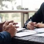 Foto de dois homens em trajes executivos sentados à mesa. À esquerda, as mãos de um homem estão entrelaçadas sobre a mesa em postura de escuta. À direita, outro homem segura uma caneta, analisando papéis com gráficos de barras e tabelas. No ambiente, há um notebook e uma varanda ao fundo com luz natural.