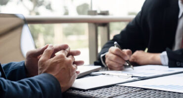 Foto de dois homens em trajes executivos sentados à mesa. À esquerda, as mãos de um homem estão entrelaçadas sobre a mesa em postura de escuta. À direita, outro homem segura uma caneta, analisando papéis com gráficos de barras e tabelas. No ambiente, há um notebook e uma varanda ao fundo com luz natural.