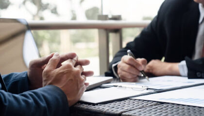 Foto de dois homens em trajes executivos sentados à mesa. À esquerda, as mãos de um homem estão entrelaçadas sobre a mesa em postura de escuta. À direita, outro homem segura uma caneta, analisando papéis com gráficos de barras e tabelas. No ambiente, há um notebook e uma varanda ao fundo com luz natural.