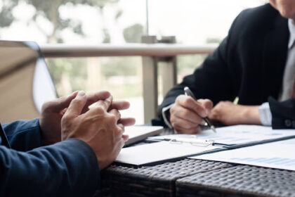 Foto de dois homens em trajes executivos sentados à mesa. À esquerda, as mãos de um homem estão entrelaçadas sobre a mesa em postura de escuta. À direita, outro homem segura uma caneta, analisando papéis com gráficos de barras e tabelas. No ambiente, há um notebook e uma varanda ao fundo com luz natural.
