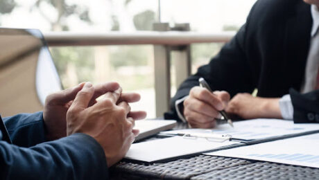 Foto de dois homens em trajes executivos sentados à mesa. À esquerda, as mãos de um homem estão entrelaçadas sobre a mesa em postura de escuta. À direita, outro homem segura uma caneta, analisando papéis com gráficos de barras e tabelas. No ambiente, há um notebook e uma varanda ao fundo com luz natural.