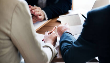 Fotografia em close-up de três profissionais em traje corporativo, sentados ao redor de uma mesa de madeira. O foco está nas mãos. Uma pessoa de terno bege à esquerda segura uma caneta dourada e aponta para um contrato papel. Uma pessoa à direita, de terno escuro, segura um bloco de notas quadriculado e uma caneta preta. Ao fundo, parcialmente visível, outra pessoa de terno escuro e um laptop. A iluminação é natural.