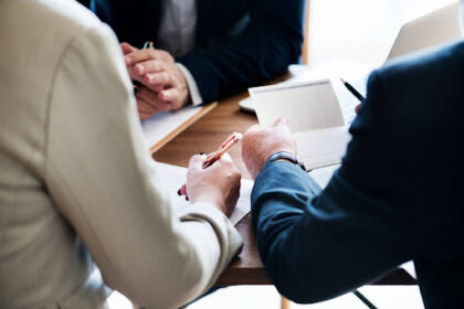 Fotografia em close-up de três profissionais em traje corporativo, sentados ao redor de uma mesa de madeira. O foco está nas mãos. Uma pessoa de terno bege à esquerda segura uma caneta dourada e aponta para um contrato papel. Uma pessoa à direita, de terno escuro, segura um bloco de notas quadriculado e uma caneta preta. Ao fundo, parcialmente visível, outra pessoa de terno escuro e um laptop. A iluminação é natural.
