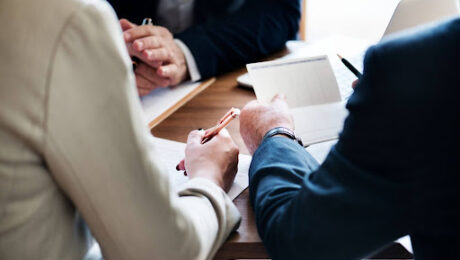 Fotografia em close-up de três profissionais em traje corporativo, sentados ao redor de uma mesa de madeira. O foco está nas mãos. Uma pessoa de terno bege à esquerda segura uma caneta dourada e aponta para um contrato papel. Uma pessoa à direita, de terno escuro, segura um bloco de notas quadriculado e uma caneta preta. Ao fundo, parcialmente visível, outra pessoa de terno escuro e um laptop. A iluminação é natural.