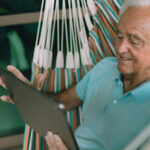 Homem idoso sorridente de cabelos brancos, vestindo camisa polo azul, sentado de forma relaxada em uma rede colorida enquanto utiliza um tablet. Ele está em uma varanda com janelas de vidro ao fundo.