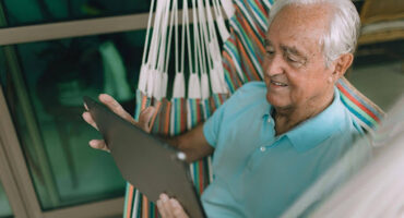 Homem idoso sorridente de cabelos brancos, vestindo camisa polo azul, sentado de forma relaxada em uma rede colorida enquanto utiliza um tablet. Ele está em uma varanda com janelas de vidro ao fundo.