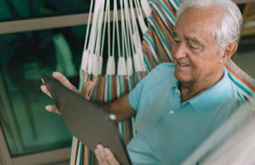 Homem idoso sorridente de cabelos brancos, vestindo camisa polo azul, sentado de forma relaxada em uma rede colorida enquanto utiliza um tablet. Ele está em uma varanda com janelas de vidro ao fundo.