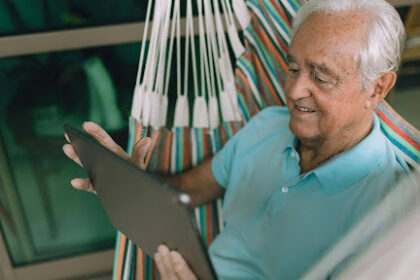 Homem idoso sorridente de cabelos brancos, vestindo camisa polo azul, sentado de forma relaxada em uma rede colorida enquanto utiliza um tablet. Ele está em uma varanda com janelas de vidro ao fundo.