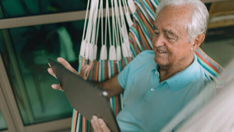 Homem idoso sorridente de cabelos brancos, vestindo camisa polo azul, sentado de forma relaxada em uma rede colorida enquanto utiliza um tablet. Ele está em uma varanda com janelas de vidro ao fundo.