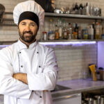 Cozinheiro sorridente com uniforme branco e chapéu de mestre-cuca em uma cozinha industrial, representando o profissional que busca a aposentadoria especial.