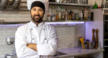 Cozinheiro sorridente com uniforme branco e chapéu de mestre-cuca em uma cozinha industrial, representando o profissional que busca a aposentadoria especial.