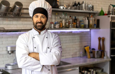 Cozinheiro sorridente com uniforme branco e chapéu de mestre-cuca em uma cozinha industrial, representando o profissional que busca a aposentadoria especial.