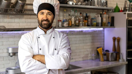 Cozinheiro sorridente com uniforme branco e chapéu de mestre-cuca em uma cozinha industrial, representando o profissional que busca a aposentadoria especial.