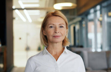 planejamento previdenciário Retrato em plano médio de uma mulher madura, loira, com expressão profissional e serena, vestindo uma camisa social branca. Ela está no centro da imagem, em pé em um escritório corporativo moderno com fundo desfocado e boa iluminação natural.