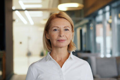 Retrato em plano médio de uma mulher madura, loira, com expressão profissional e serena, vestindo uma camisa social branca. Ela está no centro da imagem, em pé em um escritório corporativo moderno com fundo desfocado e boa iluminação natural.