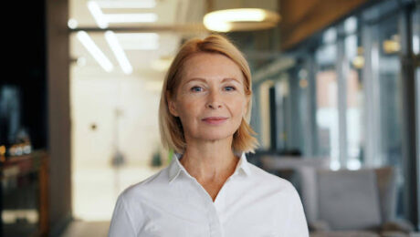Retrato em plano médio de uma mulher madura, loira, com expressão profissional e serena, vestindo uma camisa social branca. Ela está no centro da imagem, em pé em um escritório corporativo moderno com fundo desfocado e boa iluminação natural.