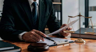 Advogado em traje formal analisando documentos sobre mesa de madeira, com balança da justiça e martelo ao lado, representando a auditoria de passivo trabalhista na sucessão de empresas.
