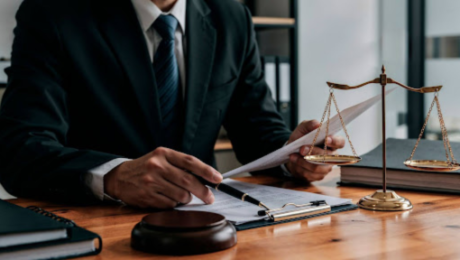 Advogado em traje formal analisando documentos sobre mesa de madeira, com balança da justiça e martelo ao lado, representando a auditoria de passivo trabalhista na sucessão de empresas.