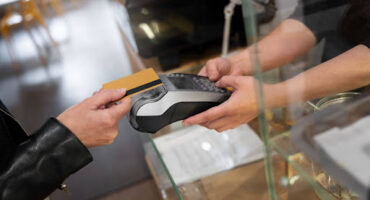 Visão em ângulo superior de uma pessoa com jaqueta preta entregando um cartão de crédito dourado para um atendente, que segura uma máquina de cartão (maquininha) preta e cinza sobre um balcão de vidro.